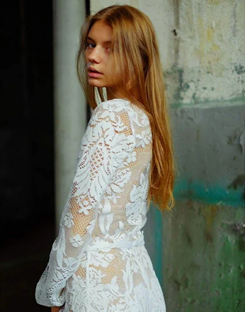 Woman wearing a white lace dress against a textured wall.