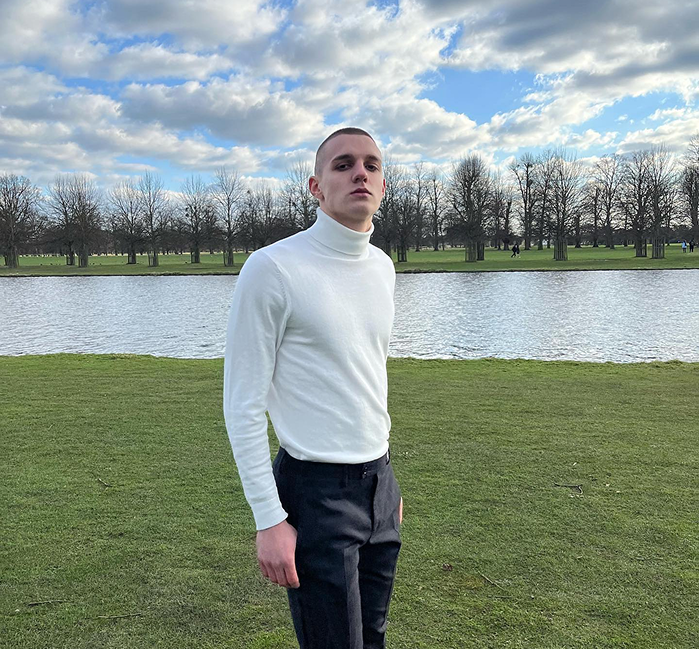 Person wearing a white turtleneck and dark pants standing in a park with a lake and trees in the background.